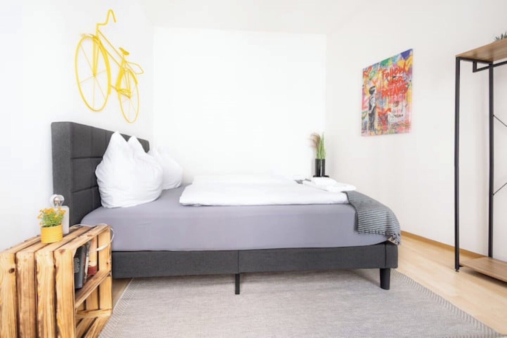 A modern bedroom with a grey bed, white pillows, a wooden nightstand, a yellow bicycle hanging on the wall, and a colorful abstract painting.