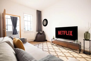 A living room with a sofa, a television displaying 'NETFLIX', a dartboard, a window with curtains, and a vase on a side table. - Ko-Living Space Opera | Suite 12 | Kitchen | Living Room | Darts | Downtown (Halle (Saale))