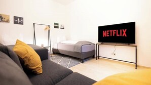 A modern living room with a grey sofa, a small bed, a standing lamp, and a TV displaying the Netflix logo.