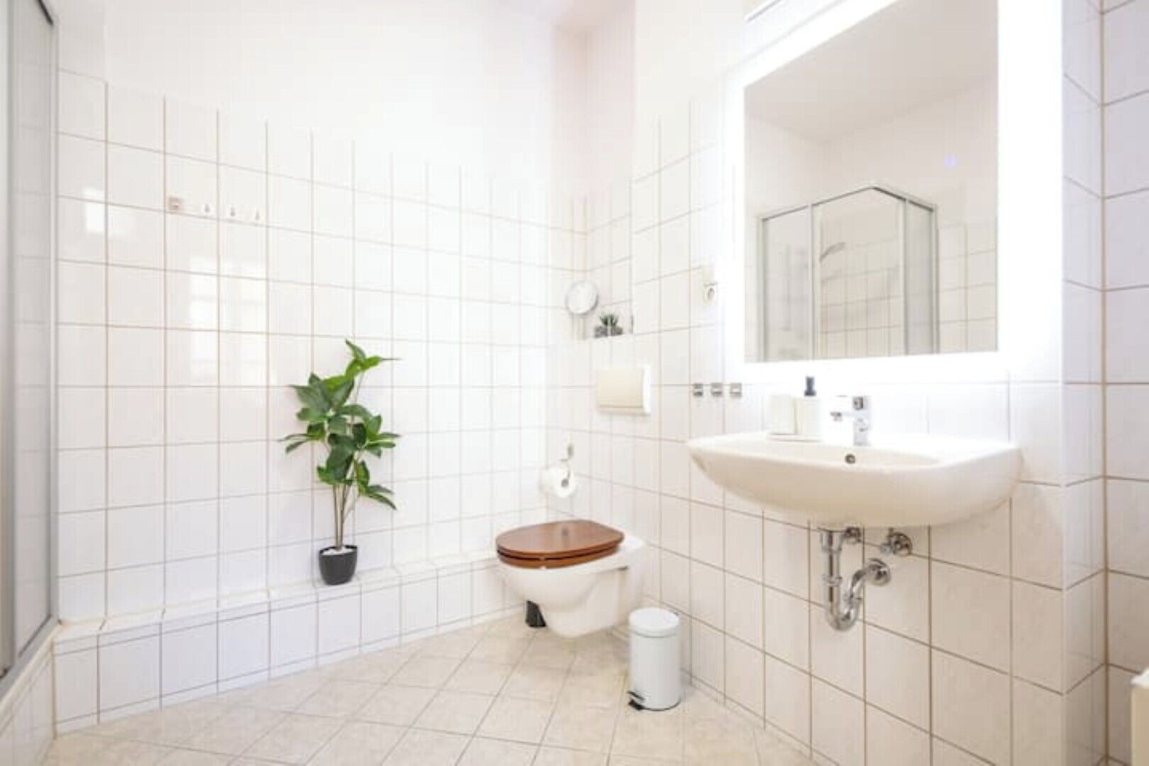 Ein Badezimmer mit weißen Fliesen, einer hölzernen Toilette, einem weißen Waschbecken und einer Topfpflanze.