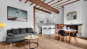 Un salon moderne avec un canapé gris, une table basse ronde et un coin repas comprenant une table et des chaises en bois. On aperçoit une cuisine à l’arrière-plan, avec des meubles blancs et une cheminée en briques.