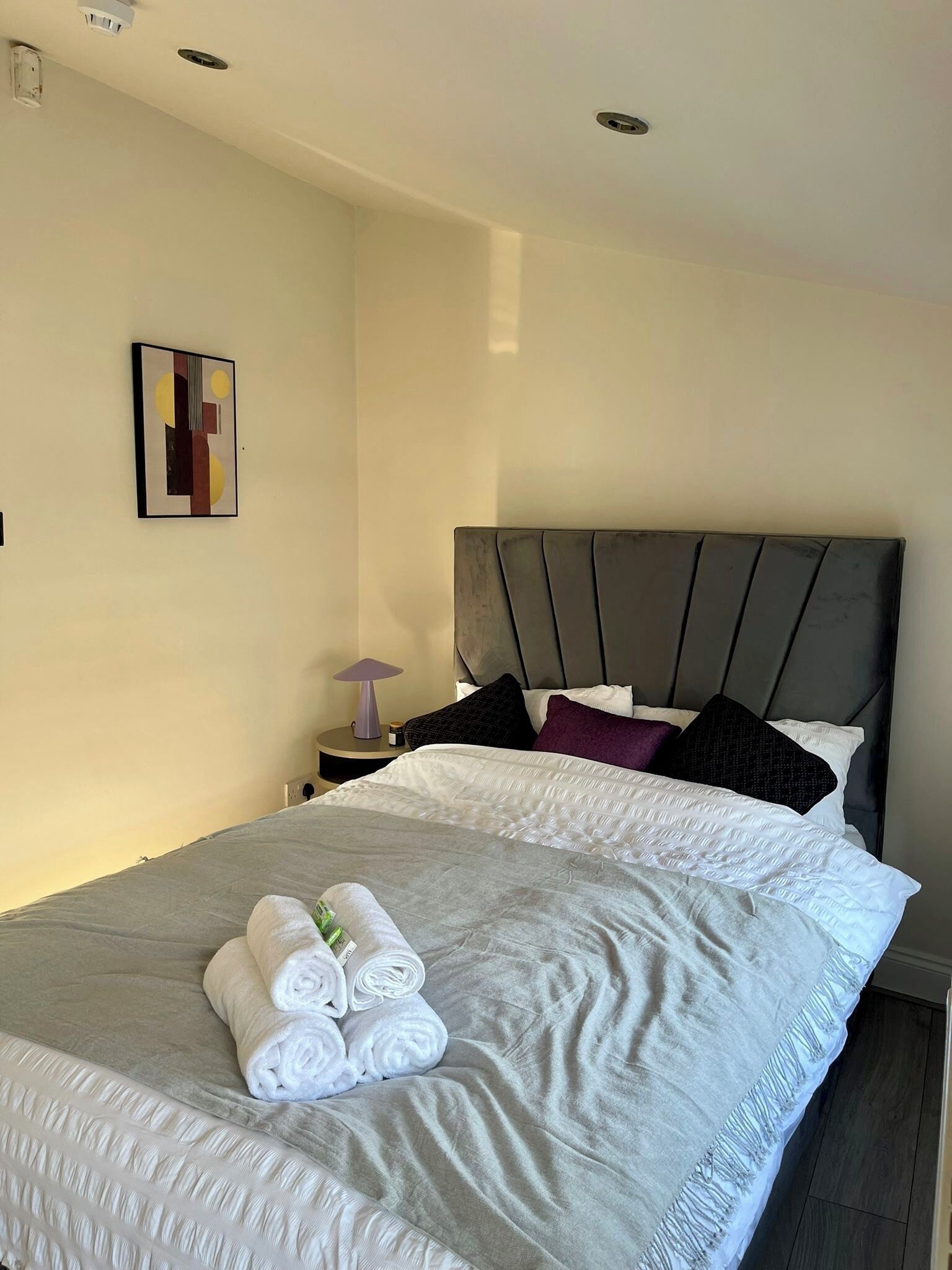Una cama bien hecha con ropa de cama blanca y gris, un cabecero y una mesita de noche pequeña con una lámpara y un adorno.