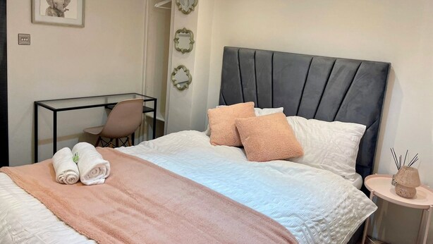 A neatly made bed with a pink blanket, two pillows, and rolled towels. There is a small bedside table with a lamp and a decorative item. A chair is placed at a desk with a mirror and a framed picture on the wall. - Cosy En-suite in The City - Shoreditch (London)