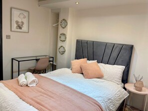 Een net opgemaakt bed met een roze deken, twee kussens en opgerolde handdoeken. Er staat een klein nachtkastje met een lamp en decoratief object. Een stoel staat bij een bureau met een spiegel en een lijst met een foto aan de muur.