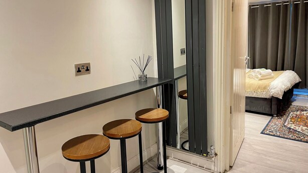 Una habitación de hotel moderna con una barra de madera oscura alargada, tres taburetes de madera, un espejo grande y una cama con cortina oscura.