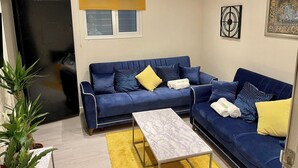 Una sala de estar con dos sofás azules, una mesa de centro blanca y cojines amarillos. Hay una ventana con una persiana amarilla, una puerta negra y una obra de arte enmarcada en la pared.