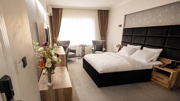 Una habitación de hotel con una cama grande, una mesita de noche de madera, un escritorio con un florero, dos sillones y una pequeña mesa con una lámpara.