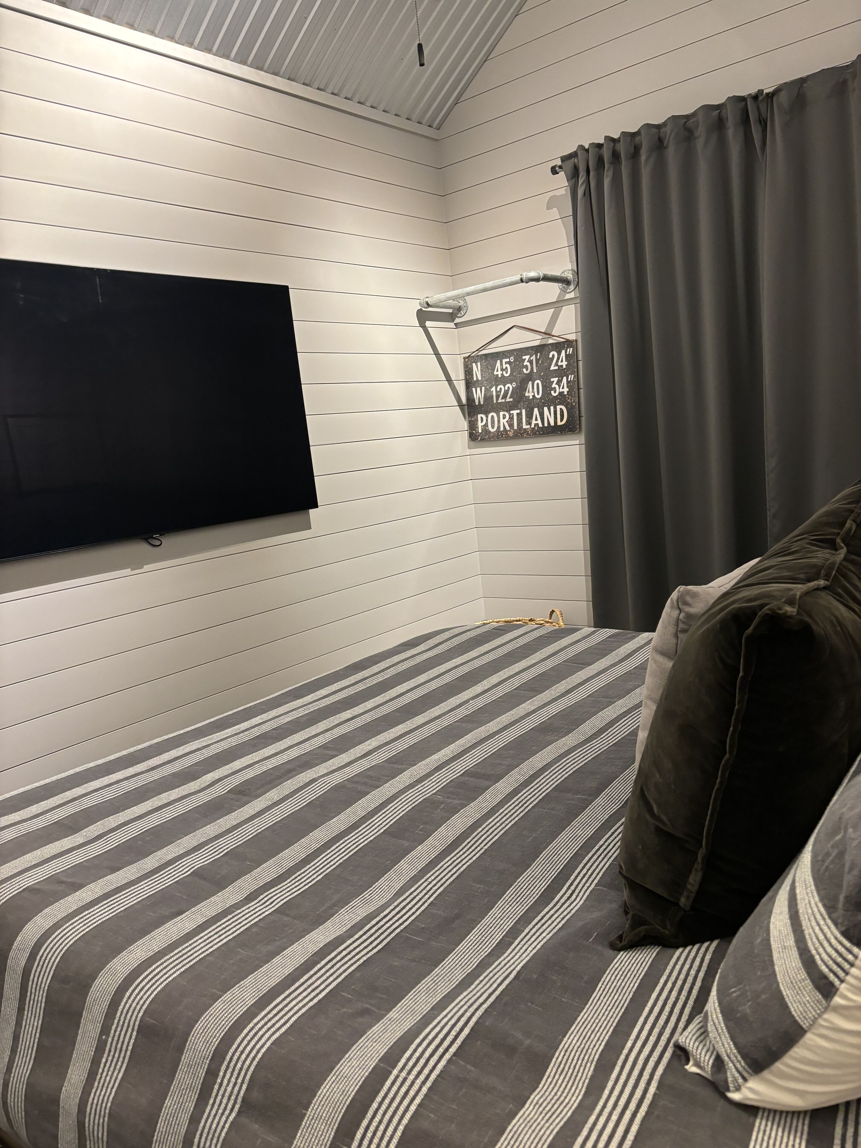 A bedroom with a bed, a mounted TV, a sign with coordinates, and a window with curtains.