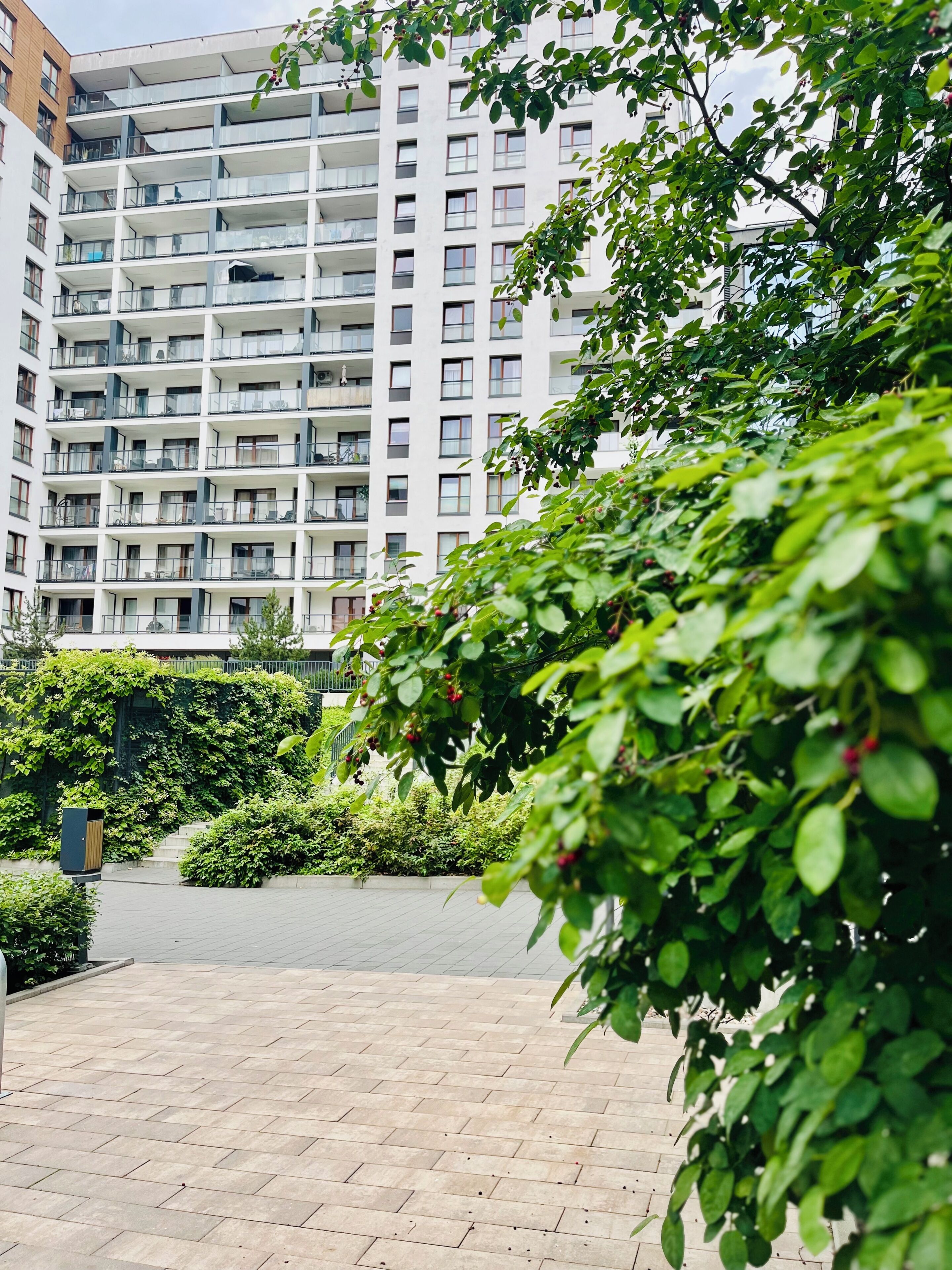 A multi-story residential building with balconies, surrounded by greenery and a paved walkway.