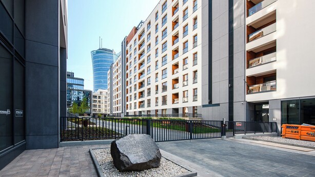 Un edificio moderno en un entorno urbano, con varios pisos, una gran escultura de roca al frente y una vista despejada de un alto edificio de cristal al fondo.