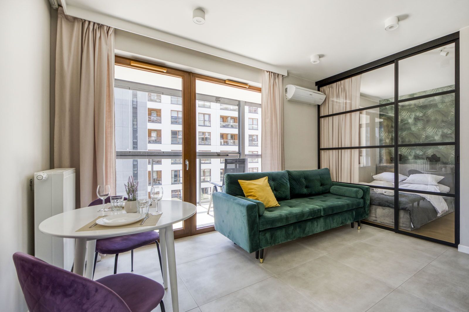 A modern living room with a green sofa, a dining table with chairs, and a large sliding glass door to a balcony.