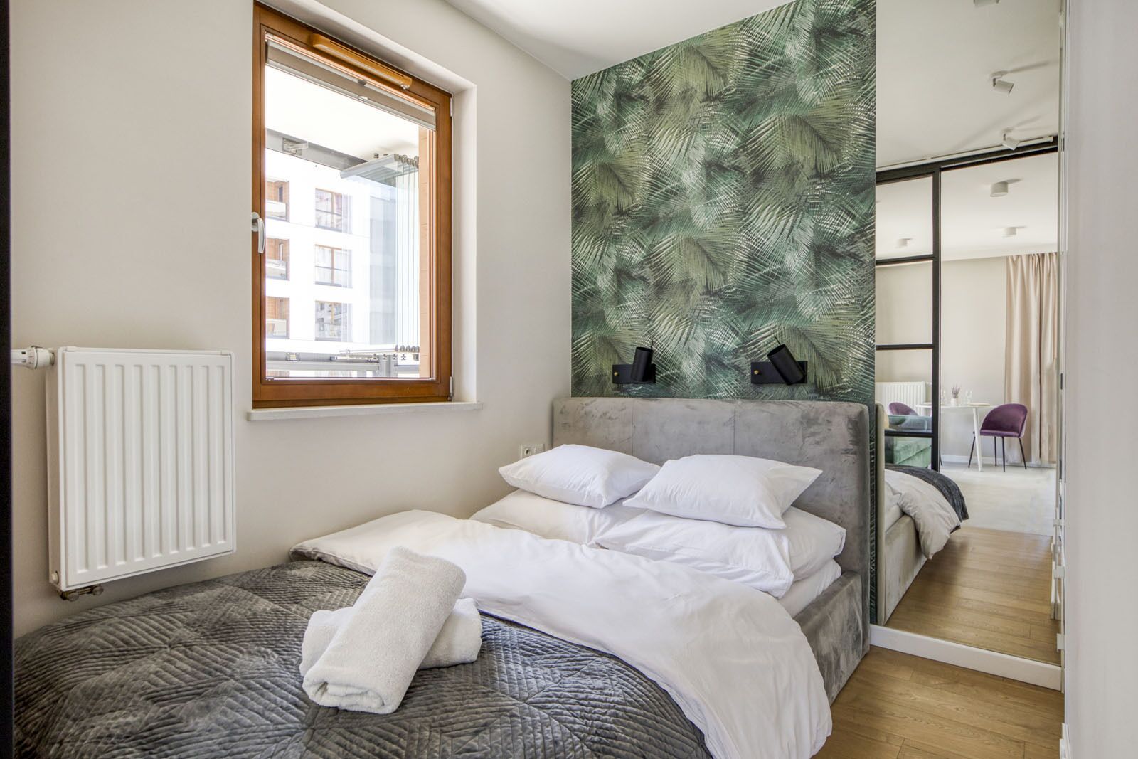 Ein Schlafzimmer mit Bett, Fenster, Heizkörper, einer Wand mit Blättermotiv und einem Spiegel, der einen Raum mit Stuhl und Tisch reflektiert.