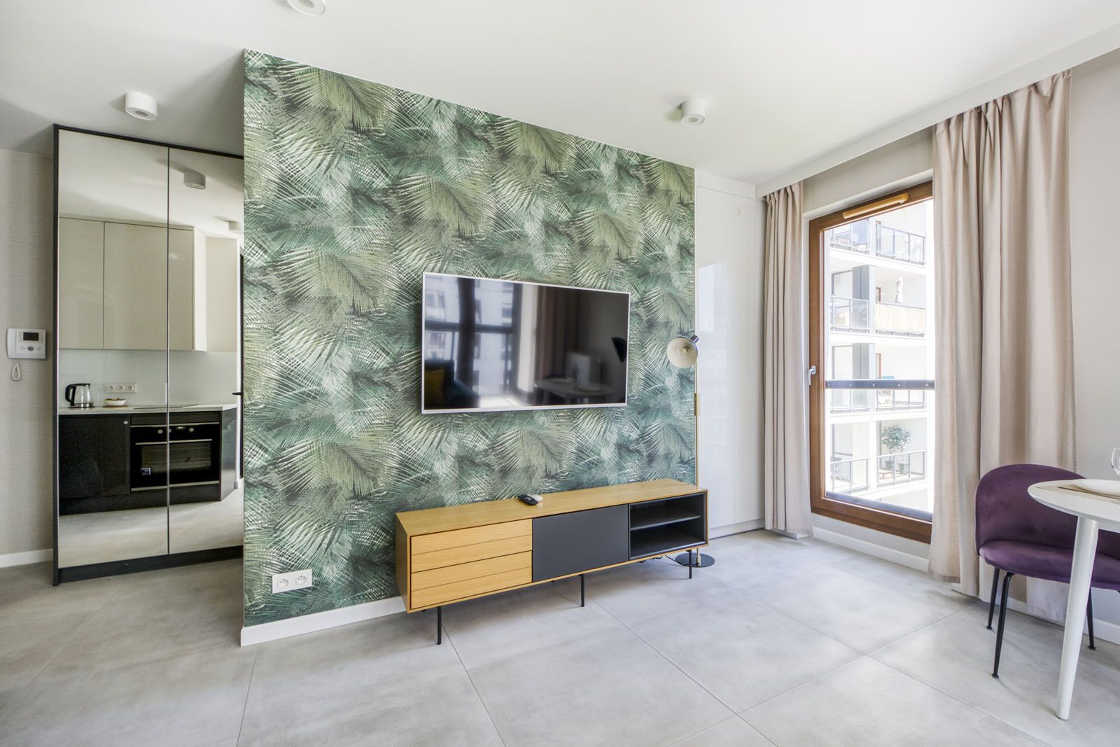 Una sala de estar moderna con un televisor de pantalla plana grande montado en una pared cubierta de un papel tapiz con motivos de hojas tropicales, un mueble de madera con cajones, una mesa de comedor con una silla morada, y una zona de cocina con horno y microondas integrados.