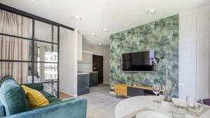 Una sala de estar moderna con un sofá, una mesa de comedor para dos, una cocina al fondo y un televisor de pantalla plana grande montado en la pared con un diseño de hojas tropicales.