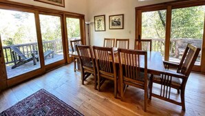 A dining area with a wooden table and chairs, large windows, and a view of the outdoors. - Forest Getaway Cozy, Inviting Cabin w Deck (Inverness)