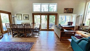 A spacious living room with a wooden floor, a dining area with wooden chairs, a blue sofa, and a large window with a view of trees. - Forest Getaway Cozy, Inviting Cabin w Deck (Inverness)