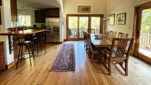 A spacious kitchen with wooden floors, a dining area with a large table and chairs, and a view through glass doors to an outdoor deck. - Forest Getaway Cozy, Inviting Cabin w Deck (Inverness)