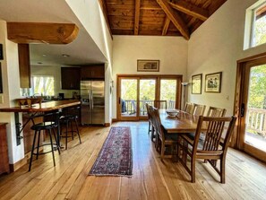 A spacious kitchen with wooden floors, a dining area with a large table and chairs, and a view through glass doors to an outdoor deck. - Forest Getaway Cozy, Inviting Cabin w Deck (Inverness)
