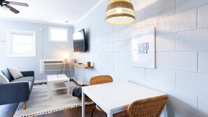 Ein modernes Wohnzimmer mit einem weißen Tisch, einem blauen Sofa, Korbstühlen, einem Deckenventilator, einem an der Wand befestigten Fernseher und einem gerahmten Zitat.