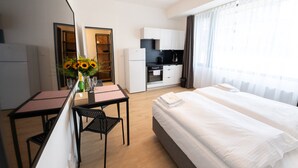 A modern hotel room with a bed, a desk with a chair, a small kitchen area, and a large window with curtains. - Post 120 Suites (Prague)