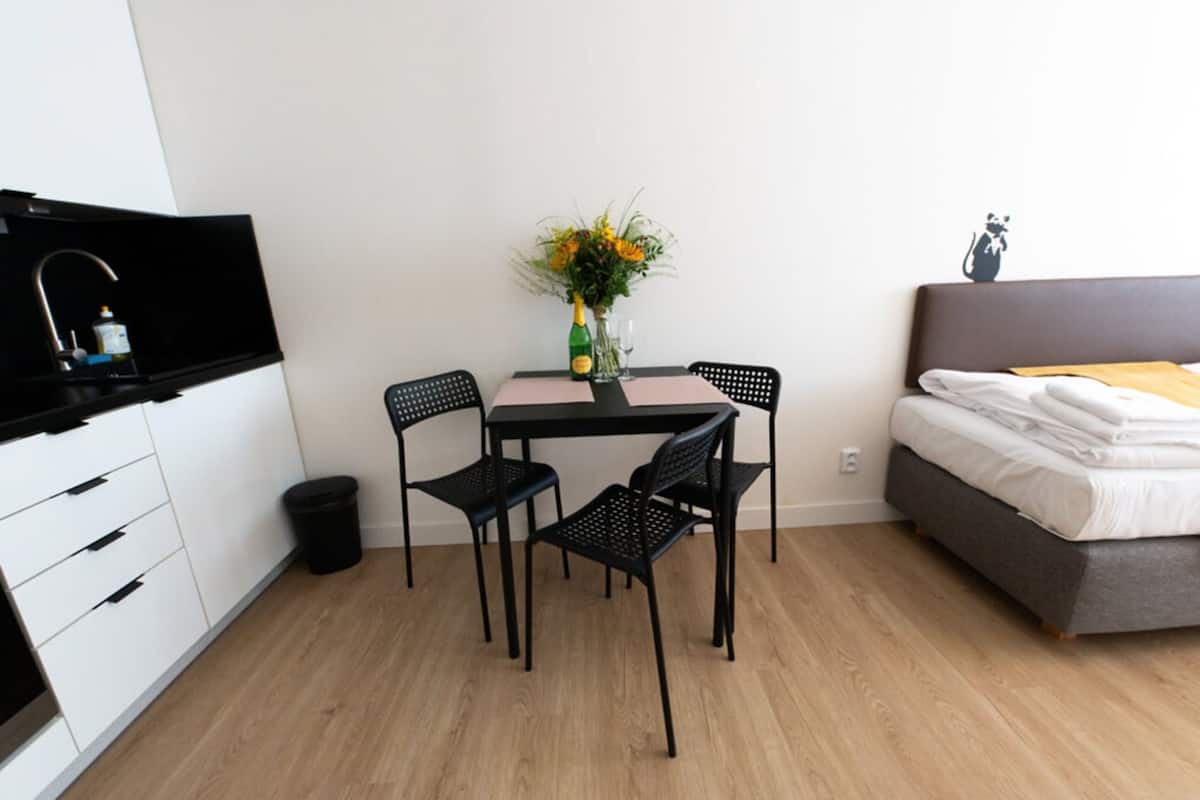 A compact bedroom with a bed, a small table with chairs, a TV, and a trash bin.