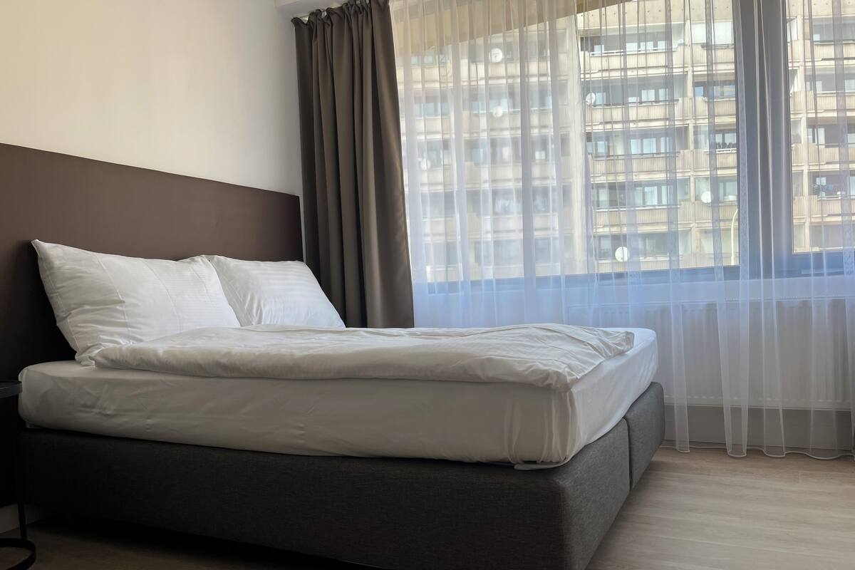 A modern hotel room with a large bed, grey walls, and a view of a cityscape through sheer curtains.
