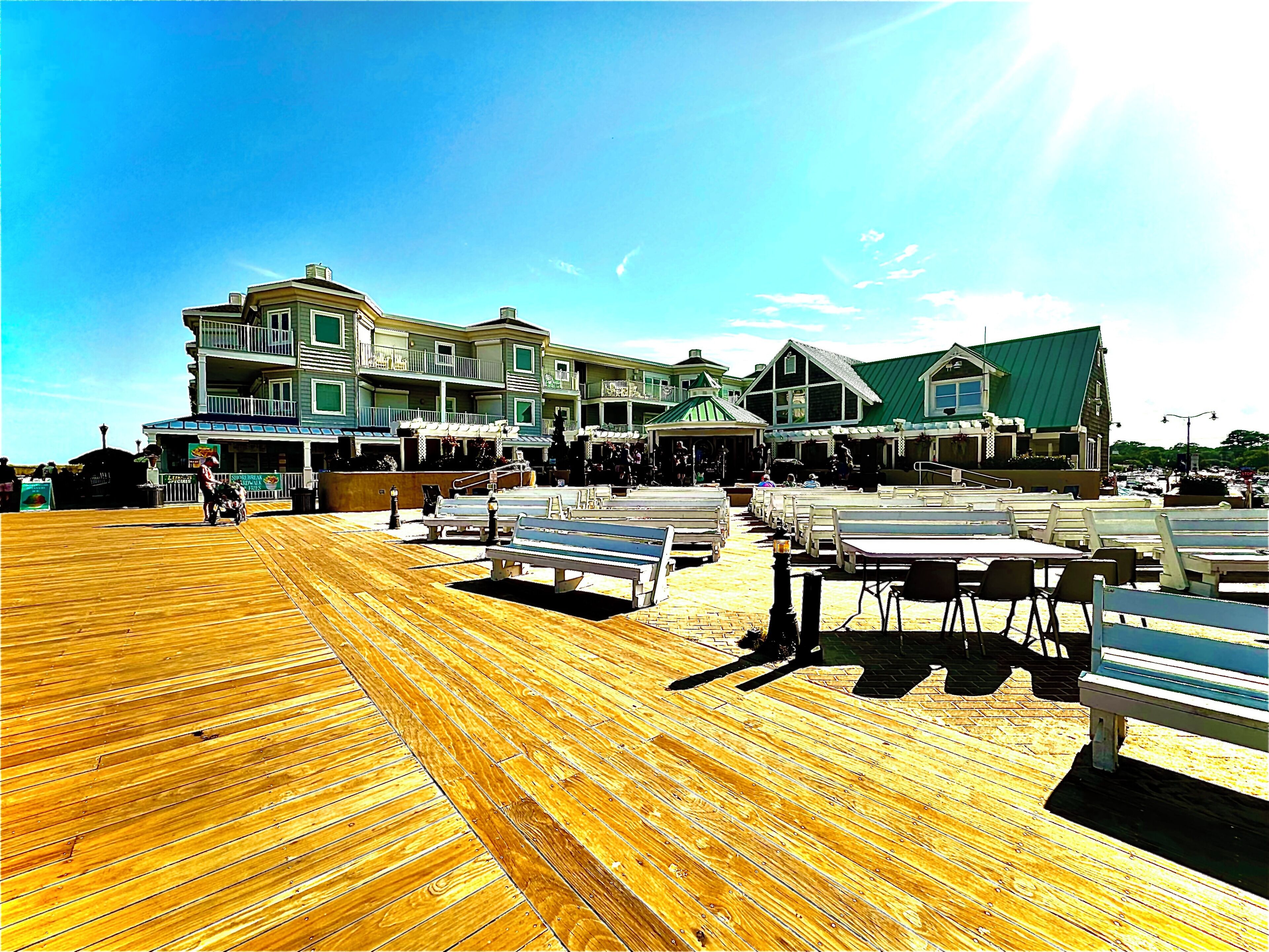 A wooden deck with tables and chairs, a building with multiple balconies, and a clear blue sky.