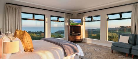 A bedroom with a large bed, a TV, a seating area, and a view of the outdoors.