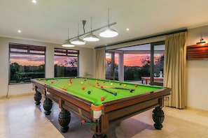 Une table de billard dans une pièce dotée de grandes fenêtres, d’un luminaire au plafond et offrant une vue sur un coucher de soleil.
