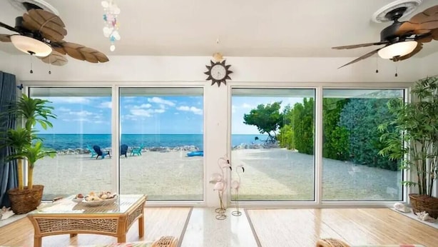 A room with large sliding glass doors leading to a beach view, a ceiling fan, a clock, and a wicker table with a decorative item.