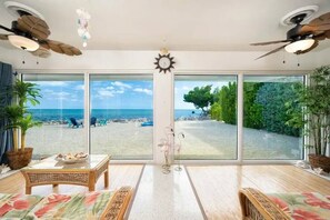 Une pièce dotée de grandes portes-fenêtres coulissantes donnant sur la plage, d’un ventilateur de plafond, d’une horloge et d’une table en osier agrémentée d’un objet décoratif.