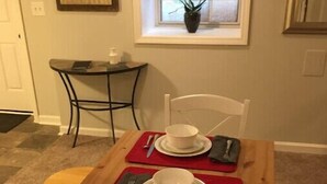 A dining area with a table set for two, a window with a plant, and a framed picture on the wall.