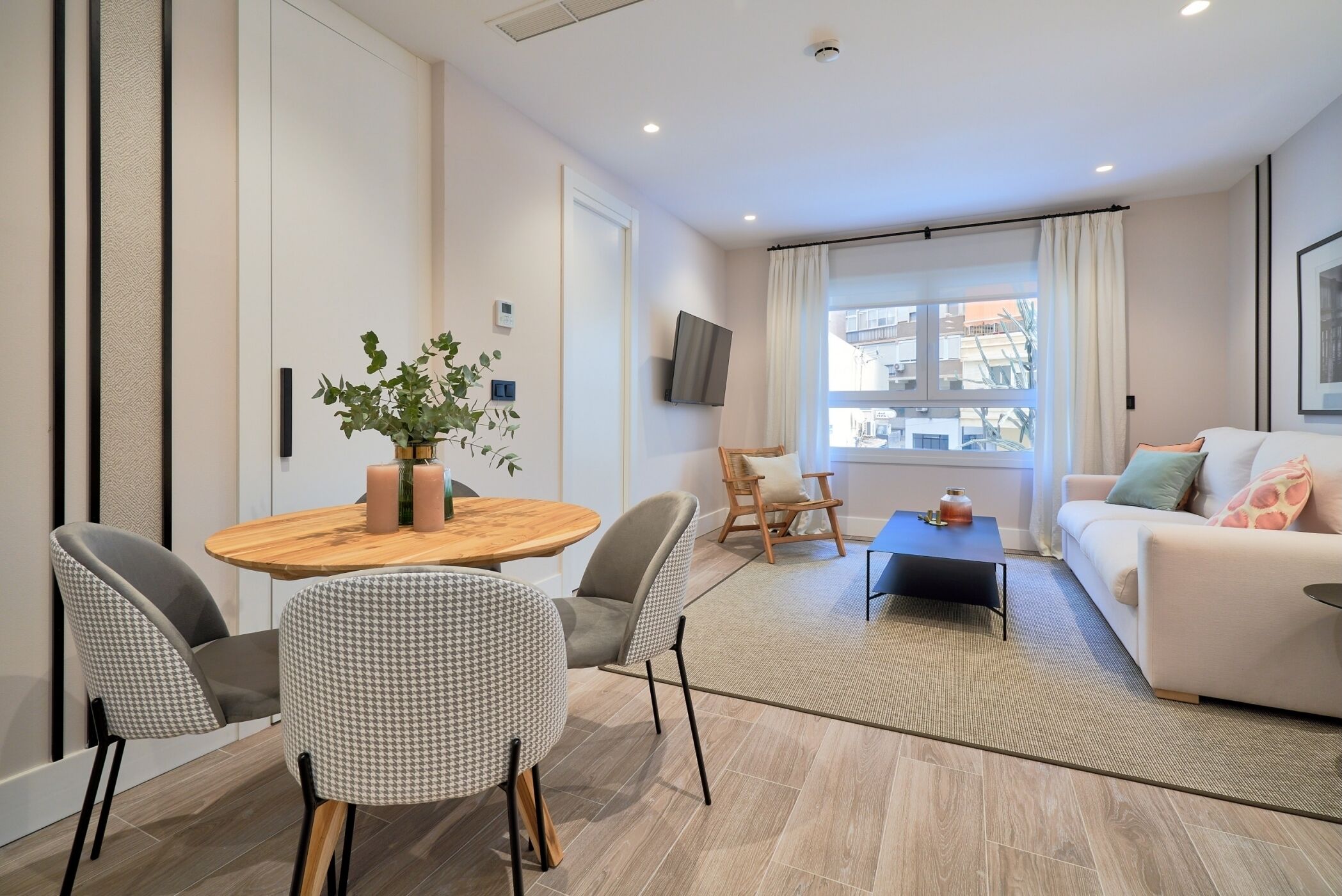 Uma sala de estar moderna com uma mesa redonda de madeira, duas cadeiras cinzas, um sofá e uma TV na parede.