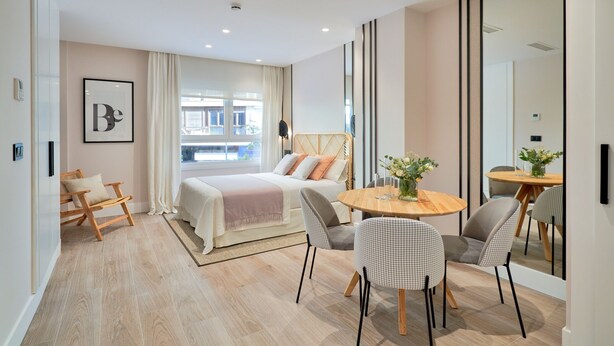 A modern hotel room with a bed, a dining table with chairs, a large mirror, and a view of the outside.