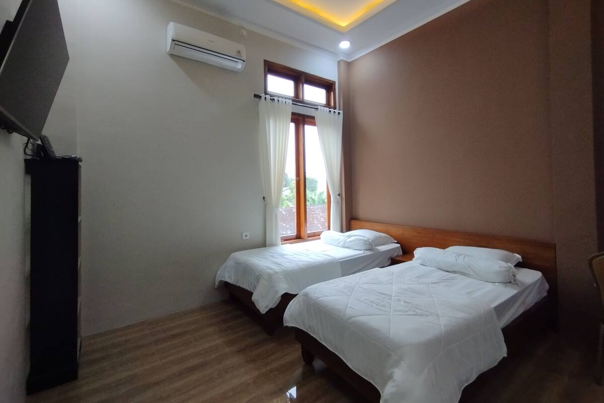 Een hotelkamer met twee bedden, een televisie aan de muur, een airconditioner en een raam met gordijnen.