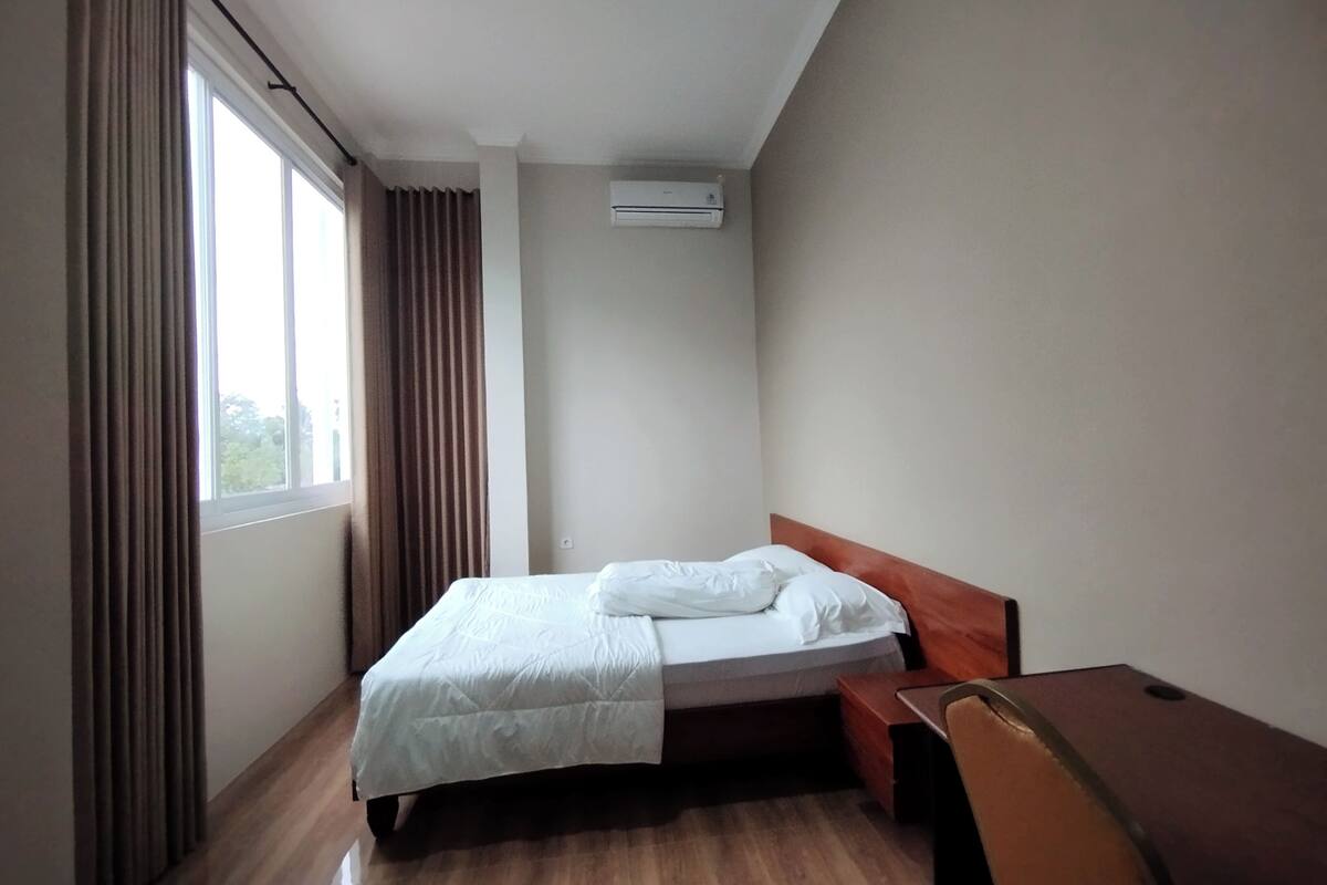 Een enkel bed met een houten hoofdeinde in een kamer met een groot raam, een airconditioning op de muur en een houten bureau.