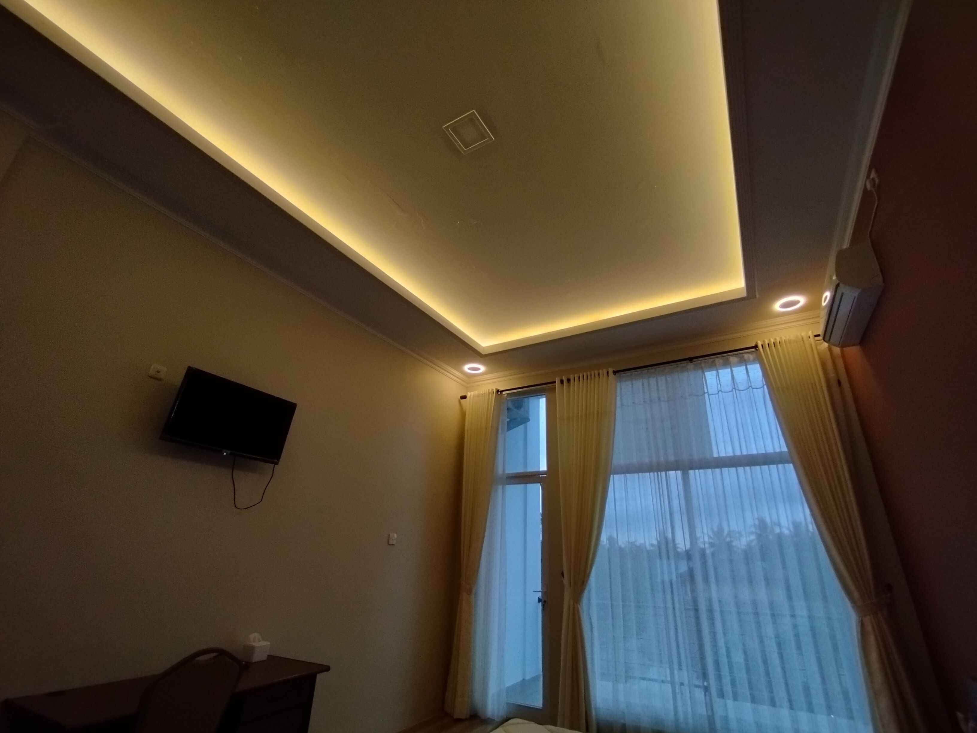 Ein Hotelzimmer mit Wandfernseher, einem Bett mit Kopfteil und einem Fenster mit durchsichtigen Vorhängen.