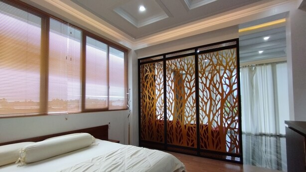 Une chambre à coucher avec un lit, un plancher en bois, de grandes fenêtres équipées de stores, un paravent décoratif et une vue sur un balcon.