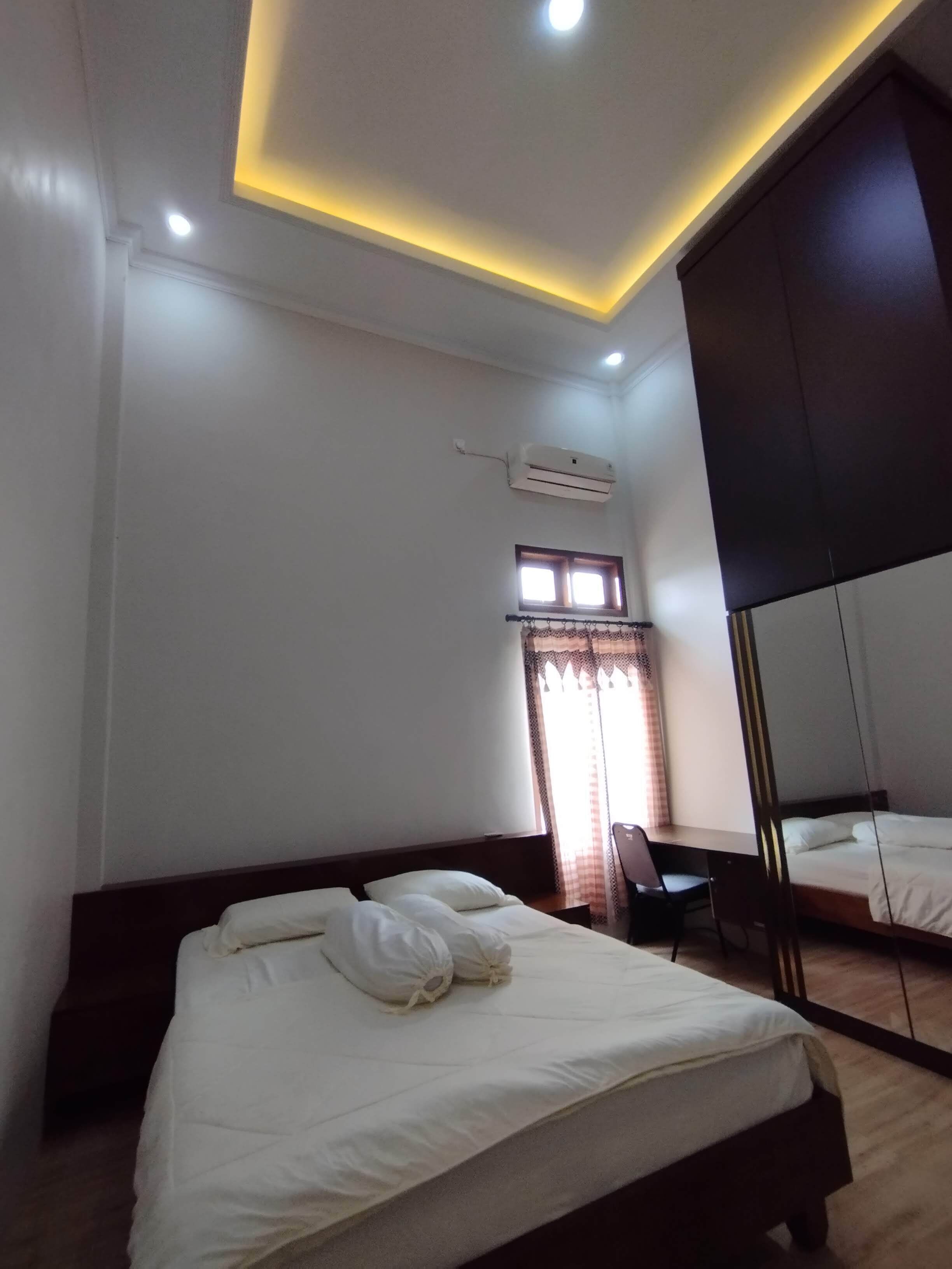 A hotel room with a bed, a desk, an air conditioning unit, a window with curtains, and a ceiling light.