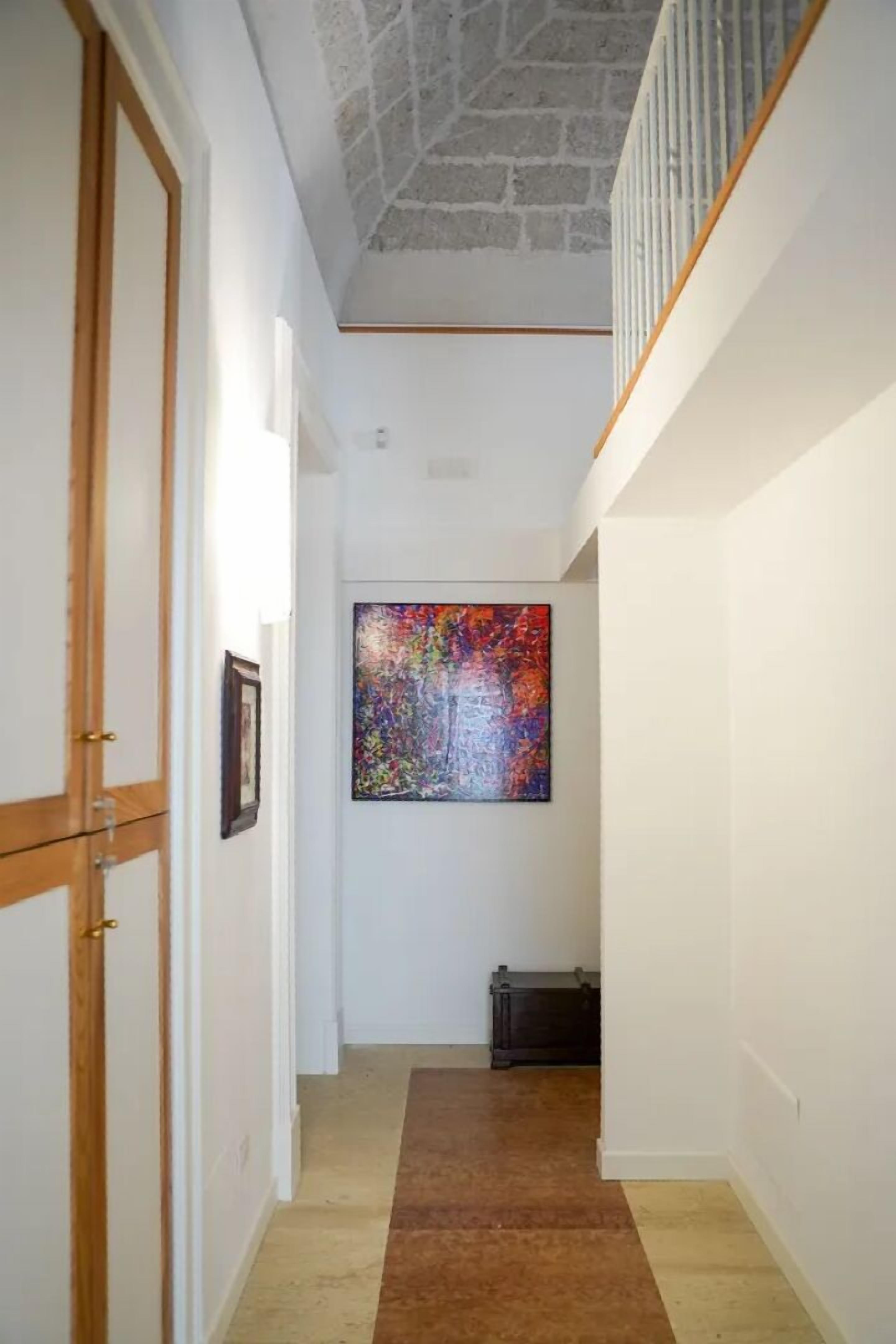 A narrow hallway with a wooden door on the left, a colorful abstract painting on the wall, and a staircase with a white railing on the right.