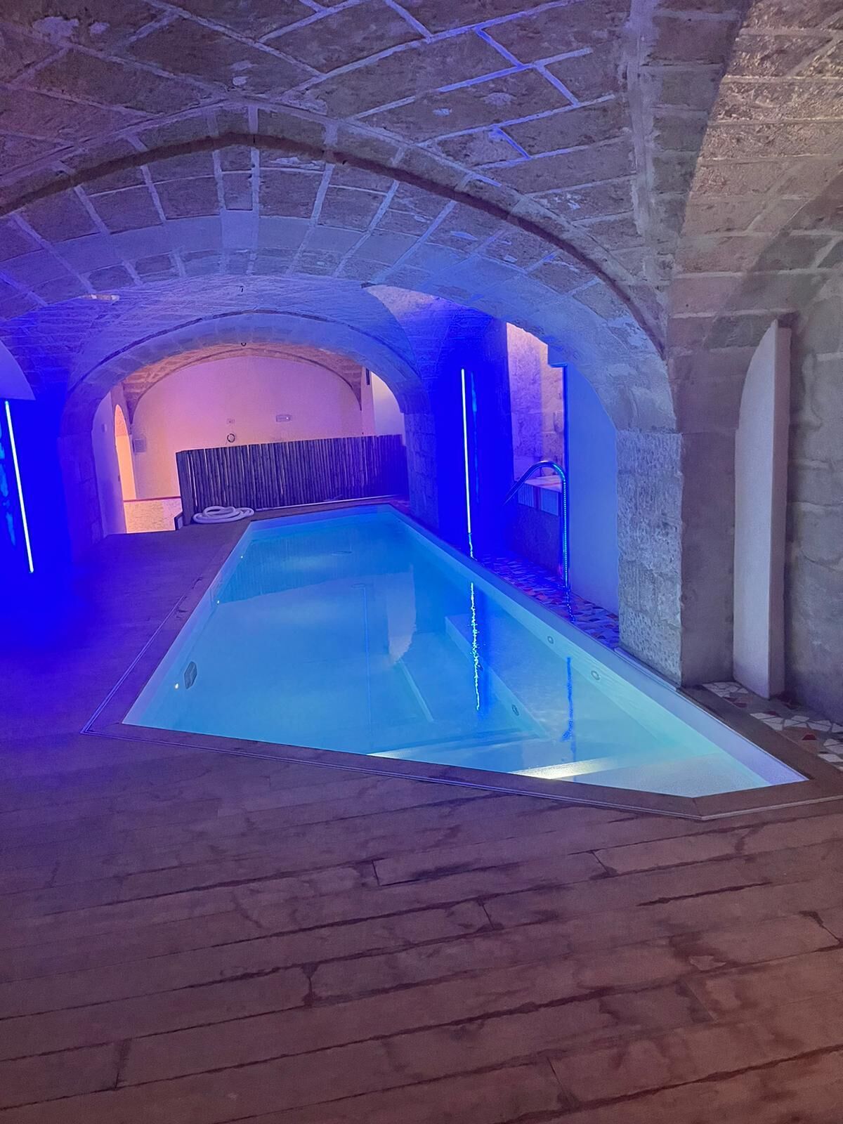 Piscina com iluminação azul, arcos de pedra e deck de madeira.