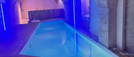 Piscina com iluminação azul, arcos de pedra e deck de madeira.