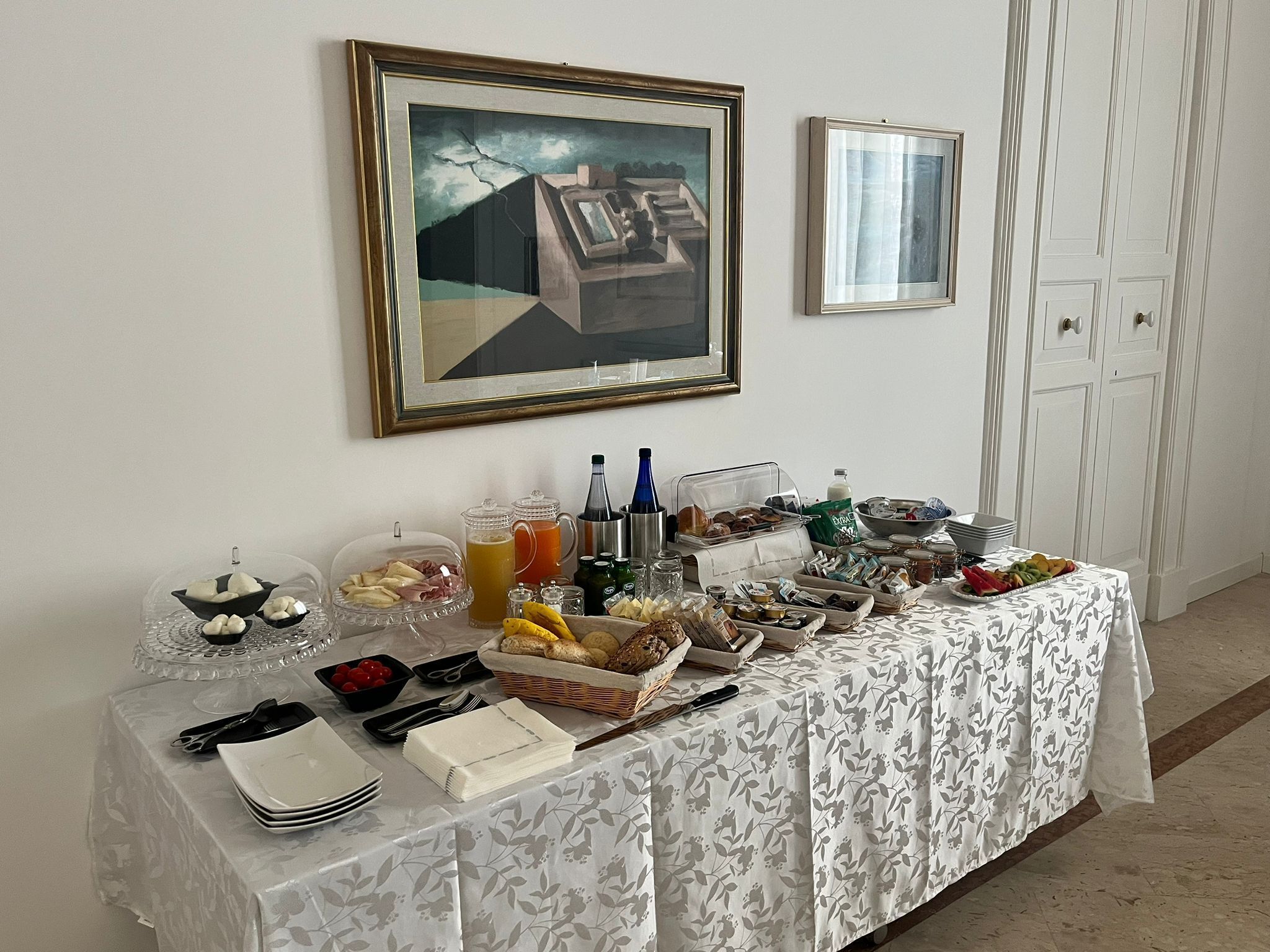 Una mesa de desayuno buffet con diversos alimentos y bebidas, una obra de arte enmarcada en la pared y una puerta blanca al fondo.