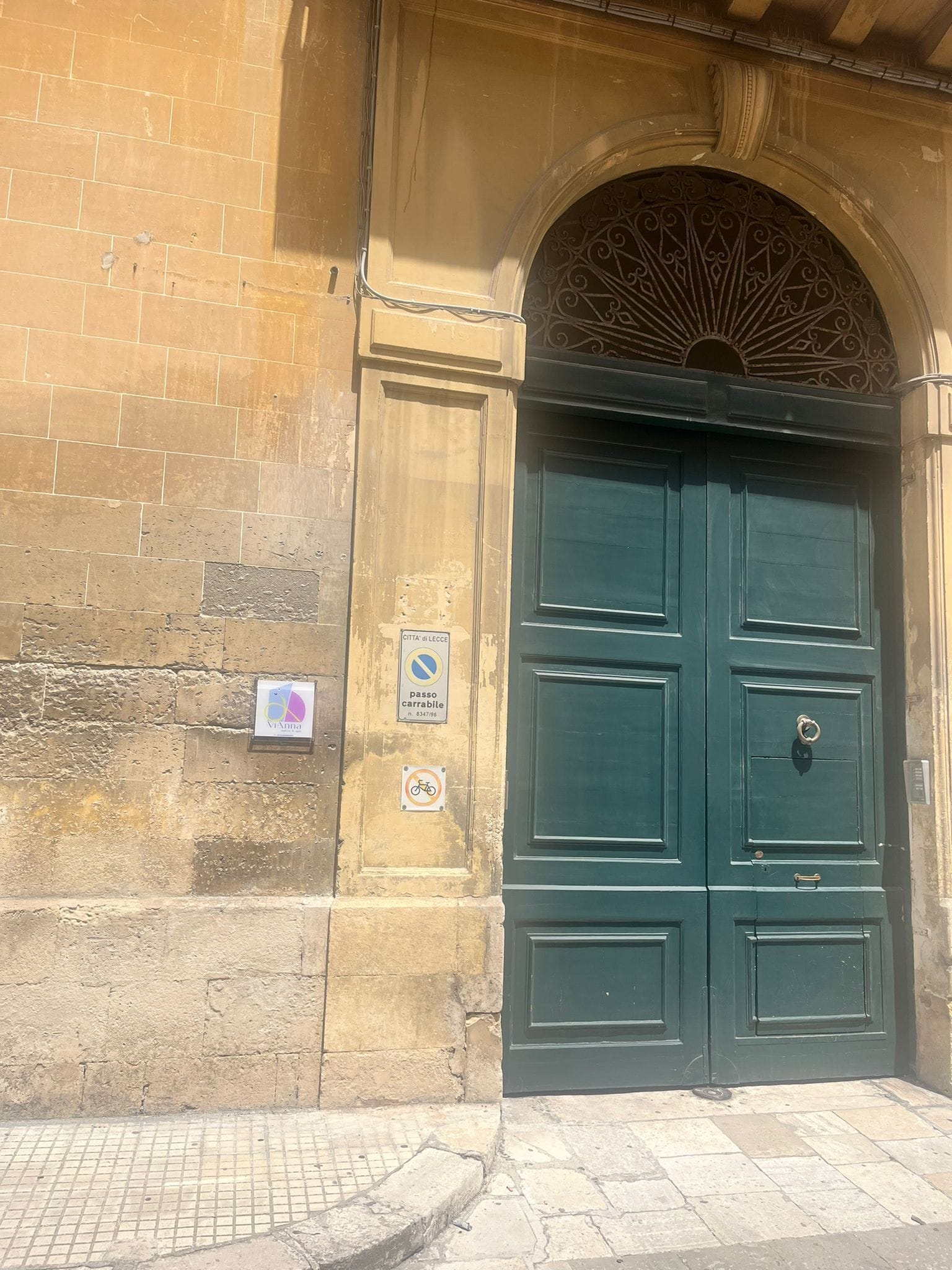 Entrada de um edifício histórico com uma grande porta verde, um muro de pedra e uma placa de metal indicando proibição de estacionamento e bicicletas.
