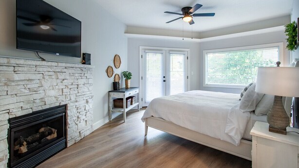 A bedroom with a stone fireplace, a bed, a ceiling fan, and a TV mounted on the wall.