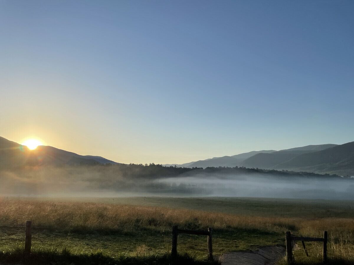 Paisaje brumoso con amanecer, una valla y colinas distantes.