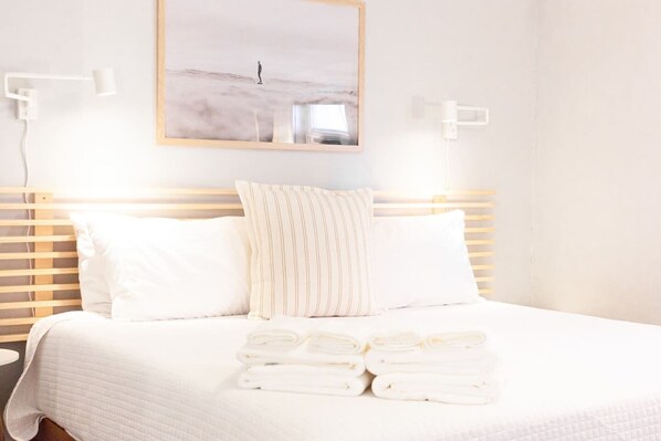 A neatly made bed with white linens and a folded towel stack, a wooden headboard, and a framed picture on the wall.