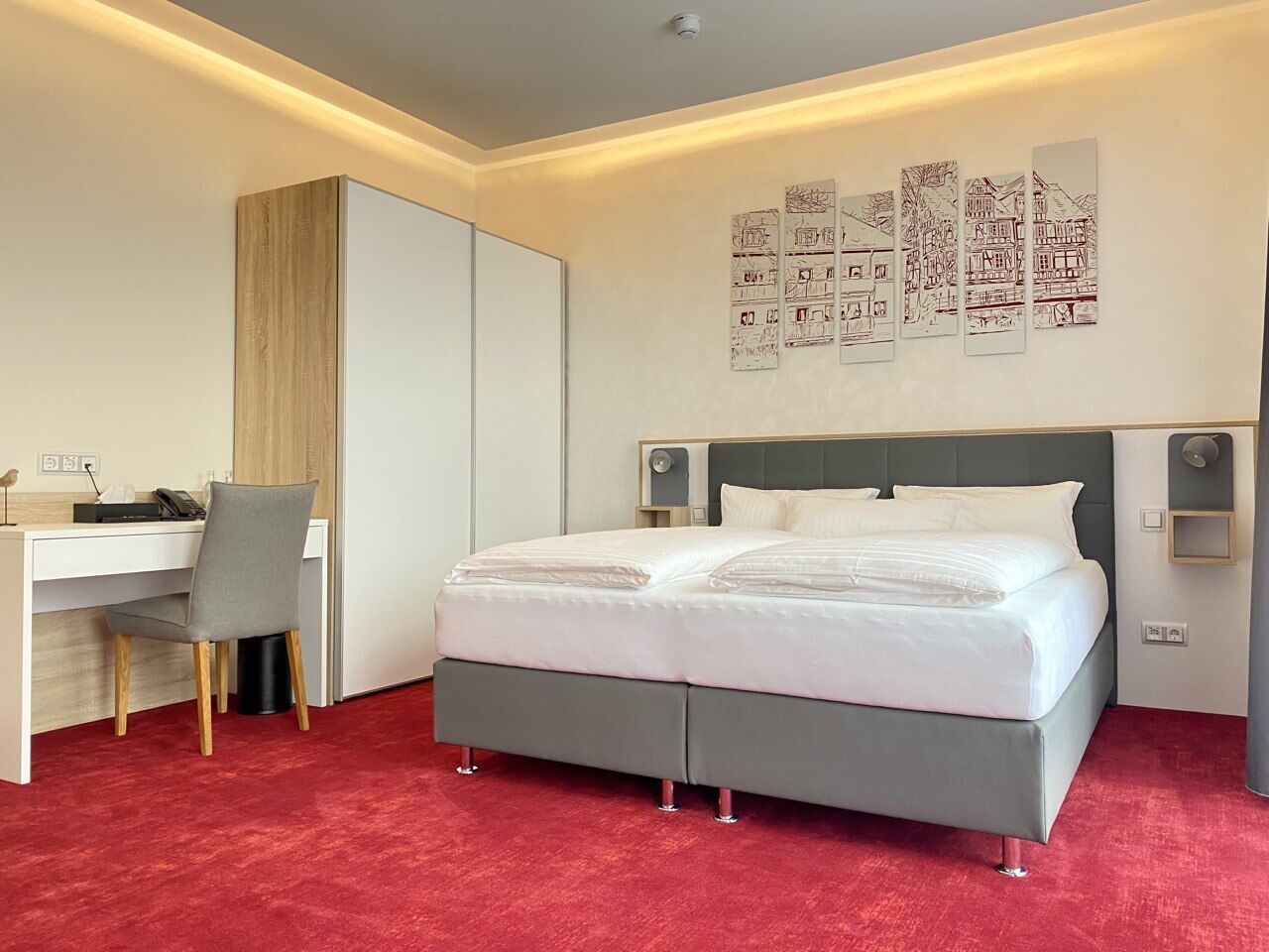 Quarto de hotel moderno com cama cinzenta, paredes brancas, tapete vermelho, escrivaninha com cadeira e obras de arte na parede.