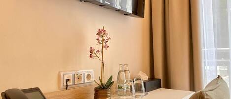 A hotel room with a flat-screen TV mounted on the wall, a desk with a telephone, a potted orchid, and a tray with glasses and water bottles.