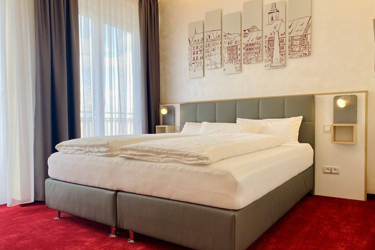 Een hotelkamer met een groot bed, een grijze hoofdeinde en een rood tapijt. Er zijn wandlampen en een raam met dunne gordijnen.